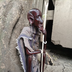 May include: A wooden sculpture of a seated figure wearing a patterned cloth. The figure has a long, thin staff in its right hand. The sculpture is carved in a dark brown color and has a detailed face.