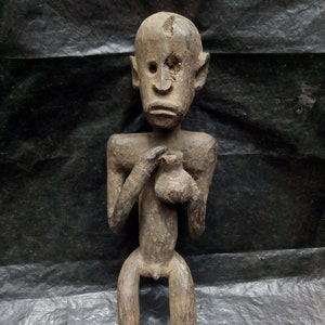 May include: A dark brown wooden sculpture of a person holding a small pot. The sculpture is carved in a stylized, abstract style. The person has a large head with a wide, flat face and a small mouth. The sculpture is about 12 inches tall.