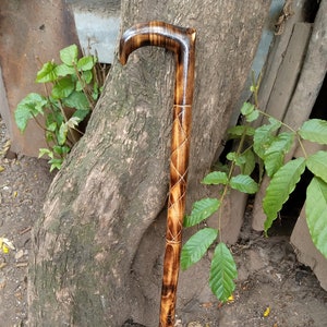 Op de afbeelding: Een houten wandelstok met een gegraveerde handgreep en een ruitvormig ontwerp. De stok is bruin en heeft een natuurlijke afwerking.