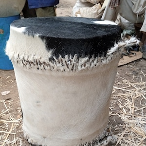 May include: A handcrafted, cylindrical stool with a cowhide exterior. The top is black and white, with a stitched seam. The sides are primarily white with black accents. The stool is approximately 18 inches tall.