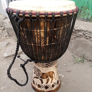 May include: A brown wooden djembe drum with a white head and black rope. The drum has a carved lion design on the bottom.