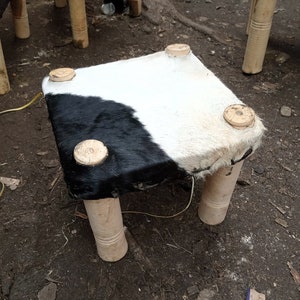 May include: A small square stool with four wooden legs. The seat is covered in black and white cowhide.