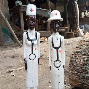 Op de afbeelding: Twee houten beeldjes van artsen, wit geschilderd met zwarte details. Beide dragen witte hoeden met rode kruisen en hebben een stethoscoop om hun nek. Een arts houdt een kleine zwarte tas.