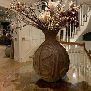 May include: A large, brown, carved ceramic vase with a wide mouth, filled with dried flowers and branches. The vase is sitting on a glass table with a wooden surface.
