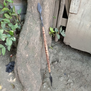 May include: A wooden spear with a black metal tip. The spear is decorated with intricate carvings and is leaning against a tree trunk.