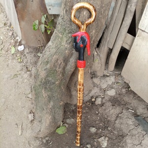 Puede incluir: Un bastón de madera con una cabeza humana tallada en la parte superior. La cabeza está pintada de negro con detalles rojos. El bastón es marrón y tiene un anillo en la parte superior.