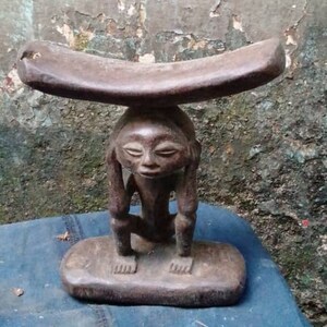 May include: A wooden sculpture of a person carrying a curved wooden seat on their head. The sculpture is dark brown and has a simple, stylized design.