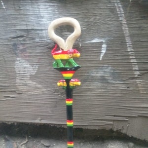 Puede incluir: Un bastón de madera con una cabeza de león tallada en la parte superior. El bastón está pintado con rayas negras, verdes, amarillas y rojas.