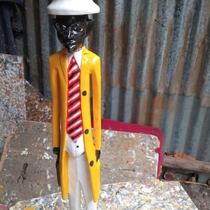 Op de afbeelding: Een houten beeldje van een persoon die een witte hoed, een geel jasje, een rood-wit gestreepte das en een witte broek draagt. Het beeldje staat op een zwarte voet.
