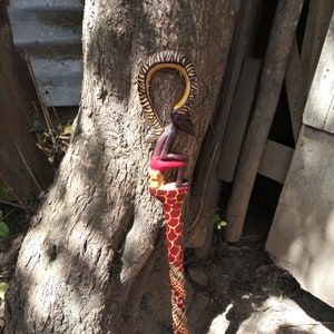 May include: A wooden walking stick with a carved figure of a person sitting on top. The stick is painted in red, yellow, and black colors.