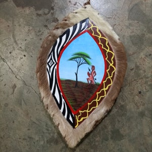 May include: A round shield made of animal hide with a painted design. The shield features a zebra pattern, a red and yellow border, and a central image of a tree and a person holding a spear. The background is blue.