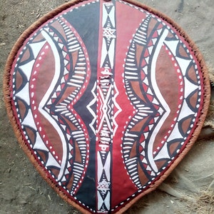 May include: A traditional African shield with a red, brown, black, and white geometric pattern. The shield is made of leather and has a woven edge.