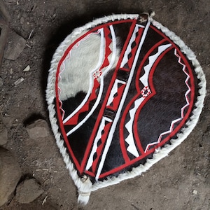 May include: A round shield with a white, black, and red geometric pattern. The shield is made of leather and has a fringe around the edge.