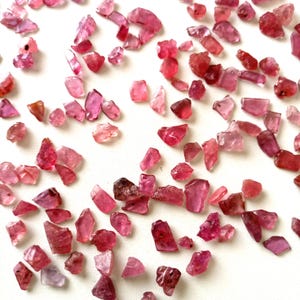 Op de afbeelding: Een verzameling ruwe, ongeslepen edelstenen in roze en rode tinten. De edelstenen variëren in grootte en vorm, met een mix van doorschijnende en ondoorzichtige stukken. De stenen zijn verspreid op een wit oppervlak.