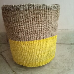 May include: A woven basket with a natural brown top and a bright yellow bottom. The basket is made of natural fibers and has a cylindrical shape.
