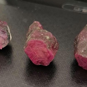 May include: Three rough, pink ruby crystals with a dark red hue. The crystals are irregularly shaped and have a natural, unpolished appearance.