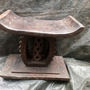 Puede incluir: Un taburete de madera marrón oscuro con un diseño tallado. El taburete tiene un asiento curvo y una base con un diseño circular.