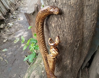 May include: A wooden walking stick carved in the shape of a snake with a smaller animal figure on top. The snake is brown and has a textured surface. The stick is leaning against a tree trunk.