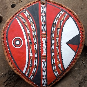 May include: A shield with a red background and geometric designs in black, white, and brown. The shield has a leather border and a loop for hanging. The design includes a central vertical pattern and a circular element.