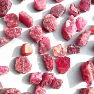 May include: A collection of raw, uncut pink gemstones scattered across a light surface. The stones vary in size and shape, displaying a range of pink hues, from light rose to deep magenta. The rough texture and natural forms of the stones are clearly visible.