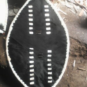 May include: A black shield with a white trim and white rectangular designs. The shield is made of leather and has a handle on the back.