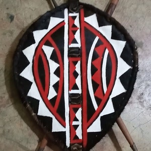 May include: A traditional African shield with a black, white, and red geometric pattern. The shield is made of leather and has a fringe around the edge.
