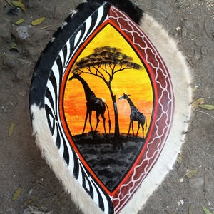 May include: A round shield with a white, black, and red zebra pattern on the outer edge. The center of the shield is painted with a yellow sunset scene with two giraffes and a tree.
