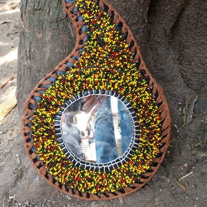 May include: A round mirror with a beaded frame in shades of brown, yellow, red, and green. The frame is made of leather and beads. The mirror is hanging on a tree trunk.