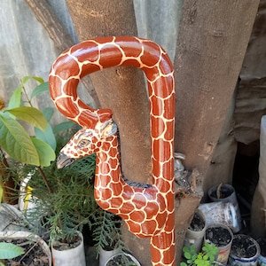 Puede incluir: Un bastón de madera tallado en forma de jirafa. La jirafa está pintada de marrón y blanco con un ojo negro. El bastón es un diseño tradicional y sería un regalo único.