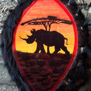 May include: A black rhinoceros silhouette painted on a wooden shield with a red border. The shield is covered in black fur and the background is a sunset with a silhouette of a tree.