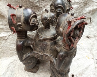 May include: A group of four dark brown wooden sculptures depicting human figures. The figures are arranged around a round pot with a lid. The figures have red fabric accents on their heads.