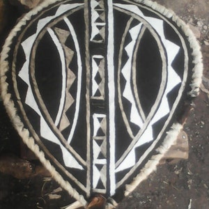 May include: A shield with a black center, white and gray geometric designs, and a fur trim. The shield has a teardrop shape and appears to be handmade. The design includes triangles, diamonds, and vertical lines.