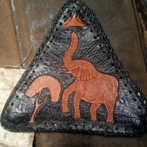 May include: A black leather motorcycle seat with a brown leather elephant and tree design. The seat is triangular in shape and has a braided edge.