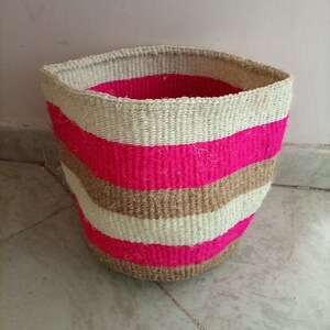Könnte beinhalten: Ein geflochtener Aufbewahrungskorb mit einem pink-braunen Streifenmuster. Der Korb ist aus Naturfasern gefertigt und hat eine zylindrische Form.