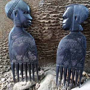 Puede incluir: Dos peines de madera negra con rostros humanos tallados y diseños intrincados. Los peines tienen una fila de dientes en la parte inferior y están hechos de madera oscura.