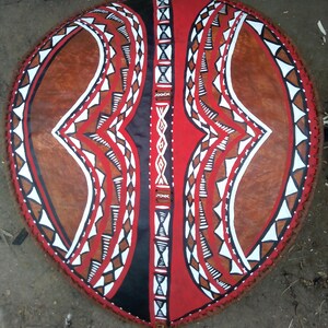 May include: A traditional shield with a red, black, and white geometric pattern. The shield is made of leather and has a fringe around the edge.