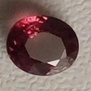 May include: A round, faceted, dark red gemstone with a slightly purplish hue.
