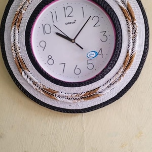 Könnte beinhalten: Eine weiße Uhr mit schwarzen Zahlen und Zeigern, umgeben von einem Perlenrahmen mit schwarzen, weißen und goldenen Akzenten. Das Zifferblatt der Uhr zeigt "OREVA".