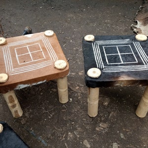 May include: Two wooden stools with a square top covered in animal hide. The tops have a grid pattern in white paint. One stool has a brown hide and the other has a black hide.