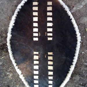 May include: A traditional African shield made of cowhide with a white fur trim. The shield has a distinctive pattern of white leather strips arranged in a symmetrical design.
