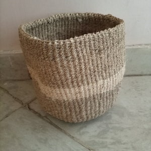 Könnte beinhalten: Ein zylindrischer Webkorb aus Naturfasern. Der Korb hat Farbtöne von Beige und Grau, mit einem helleren Band in der Mitte. Er steht auf einer grauen Oberfläche. Der Korb eignet sich zur Aufbewahrung.