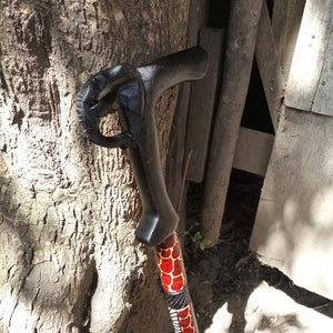 May include: A black wooden walking cane with a carved handle shaped like a fist. The shaft is decorated with a red and white pattern. The cane is leaning against a tree trunk.