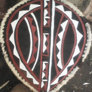 May include: A round shield with a white, red, and black geometric pattern. The shield is made of animal hide and has a fringe of white fur around the edge.