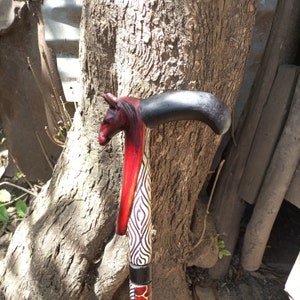 Puede incluir: Un bastón de madera con una cabeza de animal tallada en la parte superior. La cabeza está pintada de rojo y negro, y el bastón está pintado con un patrón geométrico en blanco y negro.