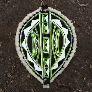 May include: A round shield with a white, black, and green geometric pattern. The shield is made of leather and has a fringe around the edge.