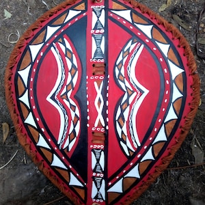 May include: A traditional shield with a red, black, and white geometric pattern. The shield is made of leather and has a fringe around the edge.