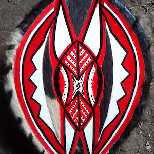 May include: A shield made of animal hide with a geometric design painted in red, white, and black. The shield is decorated with a central diamond shape and two flanking triangles.