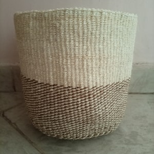 May include: A woven basket with a natural beige color and a brown band around the top. The basket is made of natural fibers and has a cylindrical shape.