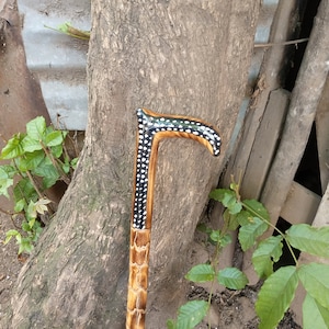 Op de afbeelding: Een houten wandelstok met een zwart-wit gepatroneerd handvat. De stok is bruin en heeft een ruwe textuur.
