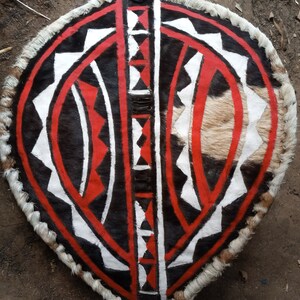 May include: A traditional African shield made of animal hide with a geometric pattern painted in black, white, and red. The shield is oval-shaped and has a fringe of fur around the edge.